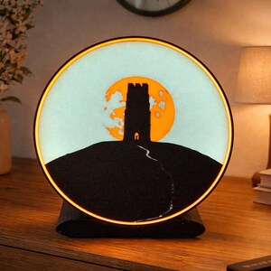 May include: A round, illuminated art piece featuring a silhouette of a tower on a hill against a yellow moon. The artwork is set in a black base, with a warm, glowing border. The scene is reminiscent of a landscape.