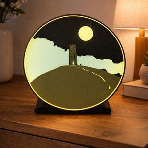 May include: A round, illuminated night light featuring a silhouette of a tower on a hill under a night sky with a full moon. The light has a black base and a warm, yellow glow, creating a cozy ambiance. It sits on a wooden surface.