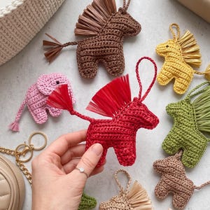 May include: A collection of handmade crochet animal keychains in various colors, including red, brown, yellow, green, and pink. The keychains feature a horse design with raffia manes and tails. A hand holds a red horse keychain.