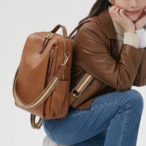 May include: A brown rectangular backpack featuring gold-tone metal zips, a top handle, and a detachable shoulder strap with a striped pattern in shades of brown, white, and tan. The bag has a smooth finish and includes side pockets.