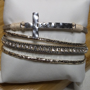 May include: A multi-strand bracelet set on a white pillow. The top strand features a silver cross with the word "BLESSED". The other strands are gold-toned and one is adorned with clear crystals. The bracelet is a religious accessory.