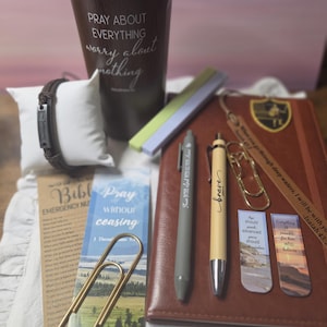 May include: A dark brown tumbler with the text PRAY ABOUT EVERYTHING worry about nothing, a brown leather-bound journal, two pens, two gold paper clips, a brown bracelet on a white pillow, and several bookmarks with religious text.