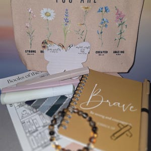 May include: A beige canvas cosmetic bag with the words "YOU ARE" and floral illustrations. A gold spiral notebook with the word "Brave" and a pen. Also, a beaded bracelet, highlighters, and a butterfly-shaped card.