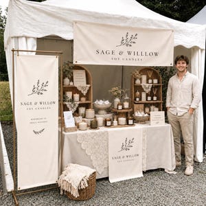 Puede incluir: Un puesto de mercado con velas de soja Sage & Willlow, pancartas color crema, manteles con encaje y estanterías de madera que contienen varias velas en frascos de vidrio, difusores de varillas y pequeños objetos decorativos.