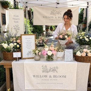 May include: A floral studio market stall featuring a linen table runner with the text Willow & Bloom Floral Studio, fresh flower bouquets in wicker baskets and glass vases, and signage listing flower varieties and bouquet sizes.