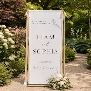 以下が含まれることがあります： 「WELCOME TO THE WEDDING OF」「LIAM and SOPHIA」「15 AUGUST 2025」「Welcome to our forever!」と黒い文字で書かれた、縦長の白いウェディングウェルカムボード。ボードはベージュのリボンが付いたゴールドのフレームから吊り下げられています。