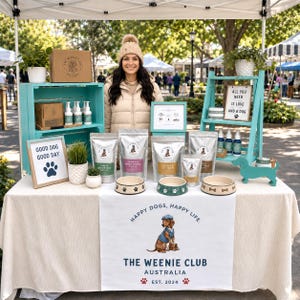以下が含まれることがあります： 白いテーブルクロスにThe Weenie Club Australiaのロゴが入ったマーケットスタンド。ドッグフードの袋、肉球模様のペット用ボウル3つ、スプレーボトル、鉢植え、そしてGood Dog Good DayやAll You Need Is Love And A Dogと書かれた看板が並んでいる。