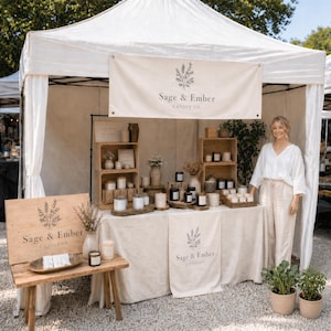 Op de afbeelding: Een kraam met kaarsen op een markt in de open lucht. De kraam heeft een houten bord en banner met de tekst "Sage & Ember Candle Co." Verschillende kaarsen in glazen potten en containers zijn gerangschikt op houten planken en een crèmekleurige tafel.