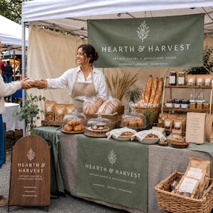 以下が含まれることがあります： Hearth & Harvestのマーケットスタンド。白い文字が書かれた緑色のバナーとテーブルランナー、ガラスドームに入ったバゲット、クロワッサン、マフィンなどの焼き菓子、木製のテーブルに並べられたパッケージ入りのグラノーラが展示されています。