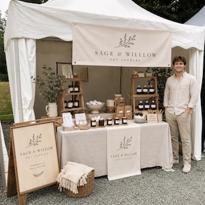 May include: A vendor booth displaying "Sage & Willow Soy Candles" products. The booth features candles, wooden shelves, and signage with the brand's logo. A person stands next to the booth, which is set up under a white tent.