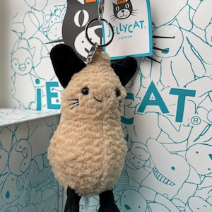 May include: A plush keychain in the shape of a small animal with a beige body, black ears, and black feet. The keychain has a silver metal ring and a blue cord. The tag reads "Jellycat London" with a black cat logo.