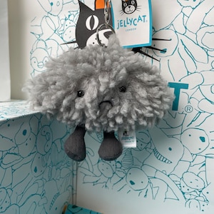 May include: A gray plush cloud keychain with a sad face and small gray feet. The keychain is attached to a blue cord and a tag with the Jellycat London logo. The background features a patterned gift bag and box.