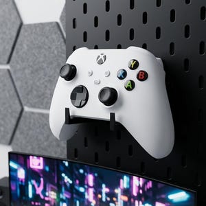 May include: A white Xbox controller mounted on a black pegboard. The controller has black joysticks and colored buttons. The text "ULTIMATE PEG-MOUNT" is visible. A curved monitor and gray hexagonal panels are in the background.