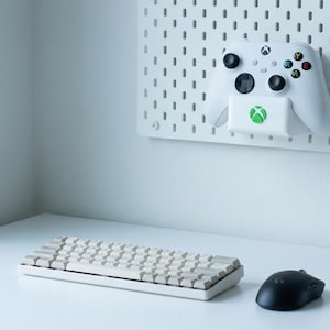 May include: A white Xbox controller with black buttons and colored action buttons is mounted on a white pegboard. A beige keyboard and black mouse sit on a white desk, suggesting a gaming setup.