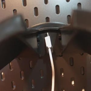 Może przedstawiać: Zbliżenie czarnej tablicy perforowanej z owalnymi otworami. Biały kabel ładujący jest podłączony do czarnego uchwytu. Kabel zwisa. Tablica perforowana jest zamontowana na ścianie.