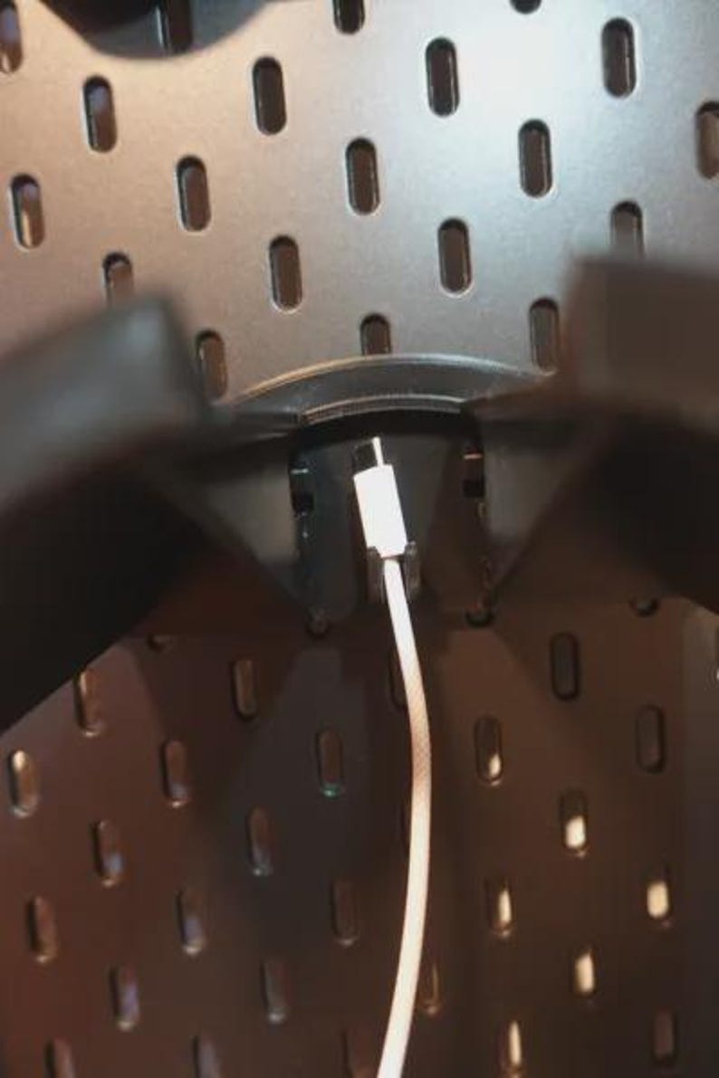 Może przedstawiać: Zbliżenie czarnej tablicy perforowanej z owalnymi otworami. Biały kabel ładujący jest podłączony do czarnego uchwytu. Kabel zwisa. Tablica perforowana jest zamontowana na ścianie.
