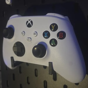 Könnte beinhalten: Ein weißer Xbox-Controller mit schwarzen Joysticks und farbigen Knöpfen (grün, rot, blau, gelb). Der Controller ist an einer schwarzen Lochwand befestigt. Das Xbox-Logo ist oben sichtbar.