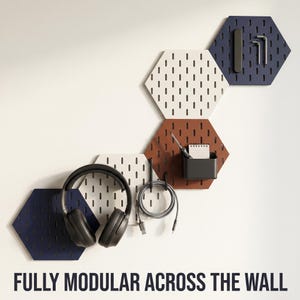 May include: Modular wall-mounted hexagon pegboards in white, brown, and navy blue. Accessories include headphones, a pen, a notepad, and tools. The text "FULLY MODULAR ACROSS THE WALL" is displayed at the bottom of the image.