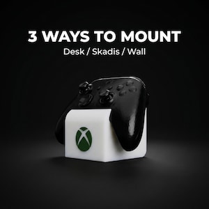 May include: A black Xbox controller rests on a white stand with a green Xbox logo. The text "3 WAYS TO MOUNT" and "Desk / Skadis / Wall" are displayed above the controller, indicating mounting options.