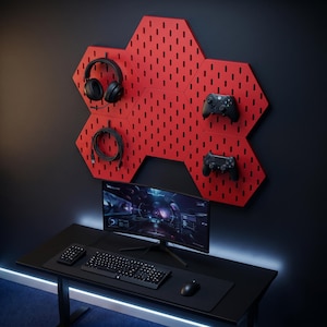 May include: A red hexagonal pegboard with gaming accessories, including headphones, controllers, and a cable. Below, a black desk holds a curved monitor, keyboard, mouse, and number pad. The setup is against a dark wall, creating a modern gaming aesthetic.