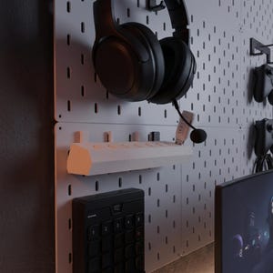 Könnte beinhalten: Gaming-Setup mit schwarzem Headset, weißem Regal und schwarzem Keypad an einer Lochwand. Das Setup umfasst ein Mikrofon und einen Computermonitor.