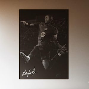 May include: A black and white poster of a soccer player in action, with a signature at the bottom. The poster is mounted on a white wall above a bed with a dark gray comforter. The room has large windows.
