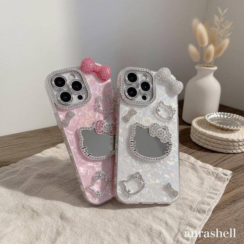 Może przedstawiać: Dwa etui na telefon, jedno r&oacute;żowe i jedno białe, ozdobione błyszczącymi kryształkami. Każde etui posiada motyw Hello Kitty z lustrzaną twarzą i akcentami w postaci kokardek. Etui mają perłowe wykończenie i są ozdobione dodatkowymi detalami z kryształk&oacute;w.