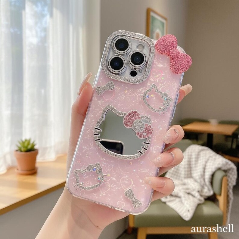 Może przedstawiać: R&oacute;żowe etui na telefon z motywem Hello Kitty. Etui ma błyszczące tło, twarz Hello Kitty z lustrem i dżetony. R&oacute;żowa kokarda jest przymocowana na g&oacute;rze.