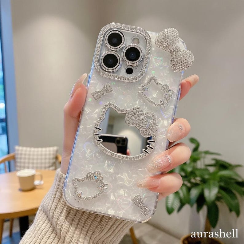 Może przedstawiać: Biało-srebrne etui na telefon z motywem Hello Kitty. Etui ma perłowe wykończenie i jest ozdobione błyszczącymi kryształkami wok&oacute;ł aparatu, twarzy Hello Kitty i kokardek. Słowo "aurashell" znajduje się w prawym dolnym rogu.