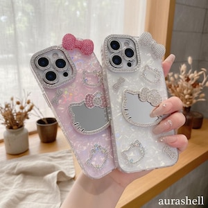 Może przedstawiać: Dwa etui na telefon, jedno r&oacute;żowe i jedno białe, każde ozdobione wzorem Hello Kitty. Etui mają błyszczące dżet&oacute;w wok&oacute;ł aparatu i twarzy Hello Kitty. Na g&oacute;rze każdego etui znajduje się kokarda.