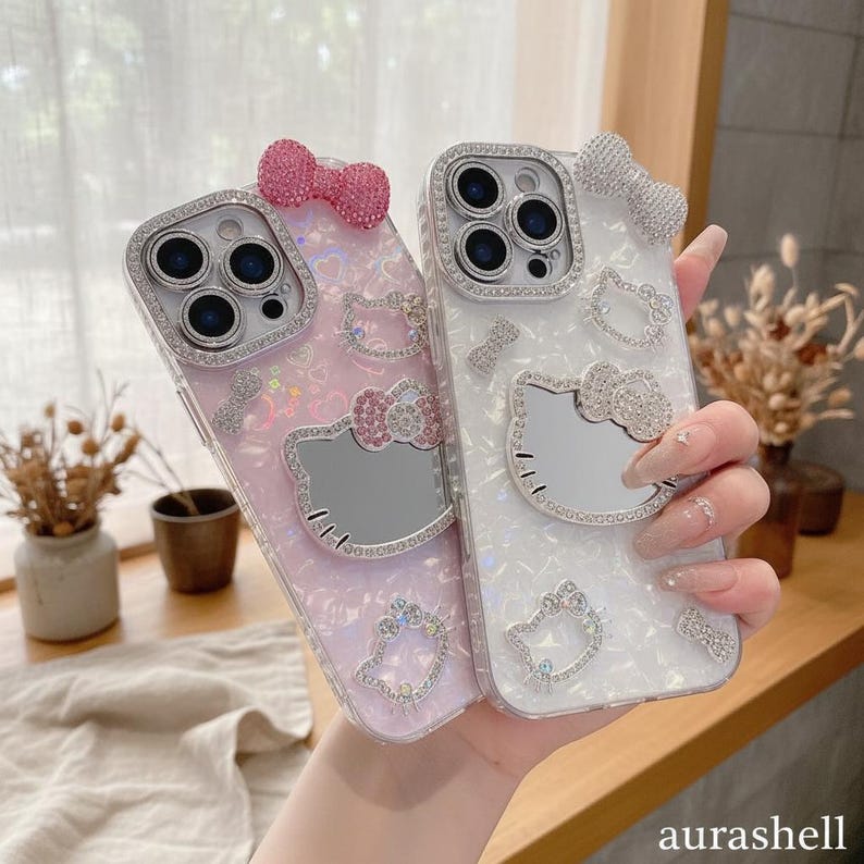Może przedstawiać: Dwa etui na telefon, jedno r&oacute;żowe i jedno białe, każde ozdobione wzorem Hello Kitty. Etui mają błyszczące dżet&oacute;w wok&oacute;ł aparatu i twarzy Hello Kitty. Na g&oacute;rze każdego etui znajduje się kokarda.