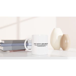 Könnte beinhalten: Weißer Keramikbecher mit gebogenem Henkel. Der Becher hat einen schwarzen Text mit der Aufschrift "NON SONO ARRABBIATO STO SOLO ANALIZZANDO IL TUO COMPORTAMENTO". Der Becher steht auf einer weißen Oberfläche mit Büchern und Vasen im Hintergrund.