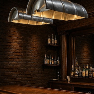 Op de afbeelding: Een donker verlichte bar met twee grote, metalen, tonvormige hanglampen. De lampen hangen boven een houten bar en een bakstenen muur. Schappen tonen diverse flessen sterke drank, wat een klassieke sfeer creëert.