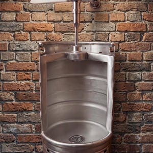 Könnte beinhalten: Ein Edelstahl-Urinal, das einem Fass nachempfunden ist, ist an einer Backsteinmauer befestigt. Das Urinal hat einen gewölbten Innenraum, einen Abfluss und ein verchromtes Spülventil. Die Backsteinmauer besteht aus roten, braunen und grauen Ziegeln.