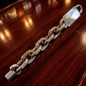 Puede incluir: Una pulsera de cadena plateada y dorada con un cierre rectangular y un lazo en el extremo. La pulsera está hecha de eslabones gruesos e interconectados y descansa sobre una superficie de madera oscura y pulida.