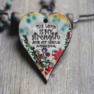 May include: Heart-shaped pendant necklace with the text "THE LORD IS MY strength AND MY SHIELD" and floral accents. The pendant is cream-colored with a brown border and a black cord.