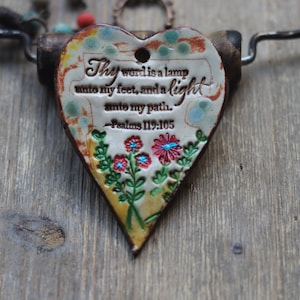 May include: Heart-shaped ceramic pendant with the text "Thy word is a lamp unto my feet, and a light unto my path. Psalms 119:105." The pendant features a floral design and is attached to a metal and leather necklace.
