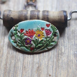May include: An oval ceramic pendant featuring a light blue glaze with a raised, hand-painted floral design. The motif includes red, yellow, and purple flowers with green stems and small red hearts. The piece has two holes on the sides for attachment.