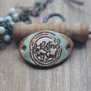 May include: Oval-shaped, distressed ceramic plaque with the inscription "He Restores my Soul" in a rustic font. The plaque has a turquoise border and a silver-colored center. A wooden handle and beaded necklace are in the background.