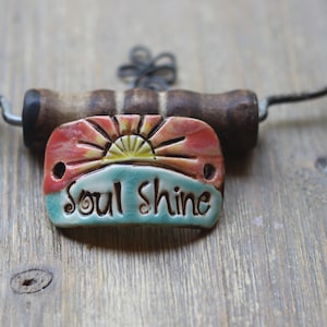 May include: A ceramic pendant with the words "Soul Shine" in cursive, featuring a sunrise design in red, yellow, and blue. The pendant is attached to a wooden corkscrew handle. A beaded necklace is visible in the background.