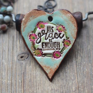 May include: Heart-shaped pendant with the text "HIS GRACE IS ENOUGH" surrounded by a floral wreath. The pendant has a turquoise and brown color scheme and is suspended from a black cord. The background is a wooden surface.