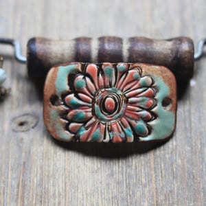 May include: A handmade ceramic pendant with a floral design in shades of coral, teal, and brown. The pendant is rectangular and attached to a wooden bar with a leather cord. Decorative beads are visible.