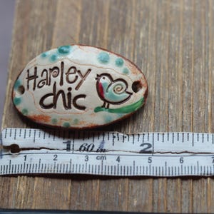 May include: A white ceramic oval tag with green and brown accents. The tag has the text "Hapley chic" and a small blue bird with a red breast. The tag is approximately 2 inches long.