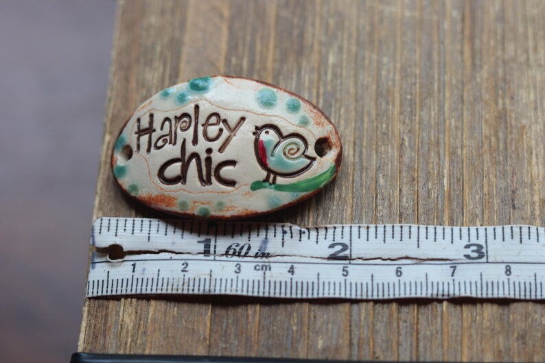 May include: A white ceramic oval tag with green and brown accents. The tag has the text "Hapley chic" and a small blue bird with a red breast. The tag is approximately 2 inches long.