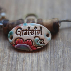 May include: An oval ceramic bead with the word Grateful embossed in black lettering. The bead features a hand-painted design of a red flower and a small blue bird. It is attached to a dark metal wire with a brown cylindrical bead and small light blue beads.