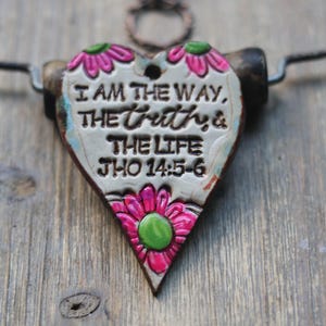 May include: A heart-shaped pendant featuring a cream-colored center with the embossed black text I AM THE WAY, THE TRUTH & THE LIFE JHO 14:5-6. The pendant is decorated with bright pink and green floral accents and hangs from a dark cord necklace.