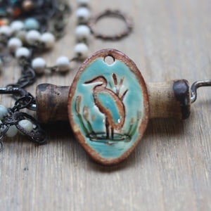 May include: An oval ceramic pendant featuring a hand-painted teal heron standing in water with reeds. The pendant has a rustic brown glazed edge and a small hole at the top for hanging. It is displayed alongside a beaded necklace with white and blue beads.