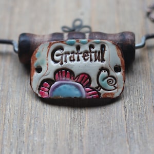 May include: A white ceramic pendant with the word "Grateful" in brown lettering. The pendant is decorated with a colorful flower and a bird design. The pendant is attached to a brown cord.