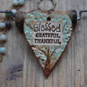 May include: A heart-shaped ceramic pendant with a light green and brown glazed finish, featuring the embossed text Blessed, GRATEFUL, THANKFUL. The pendant hangs from a dark metal wire necklace adorned with small, round, light blue beads on a chain.