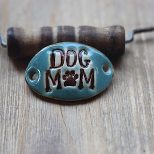 May include: A teal ceramic dog tag with the words "DOG MOM" and a paw print. The tag is oval-shaped with two holes and is set against a wooden background. A wooden handle is visible in the background.